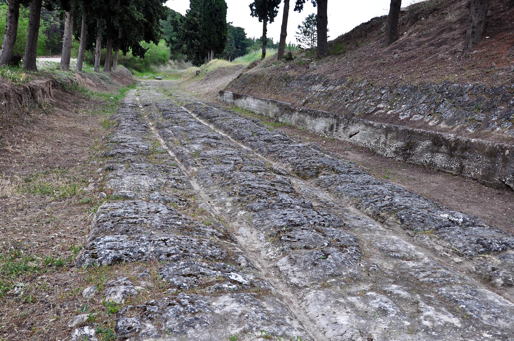 上图：哥林多铺石路（Corinth Diolkos）。哥林多城位于哥林多地峡（Isthmus of Corinth）西南方，这地峡连接希腊本土南部和伯罗奔尼撒半岛，西北有爱奥尼亚海（Ionian Sea）哥林多湾（Gulf of Corinth）的列该翁港（Lechaion），东南有爱琴海（Aegean Sea）撒罗尼克湾（Saronic Gulf）的坚革哩港（Cenchraea or Kechries）。在哥林多地峡运河开通之前，两湾之间的客旅与货物往来都经由这条铺石路中转。哥林多城以地利之便，坐收南北和东西方贸易路线上的各种税赋，所以被形容为「富裕之城」（wealthy city）。